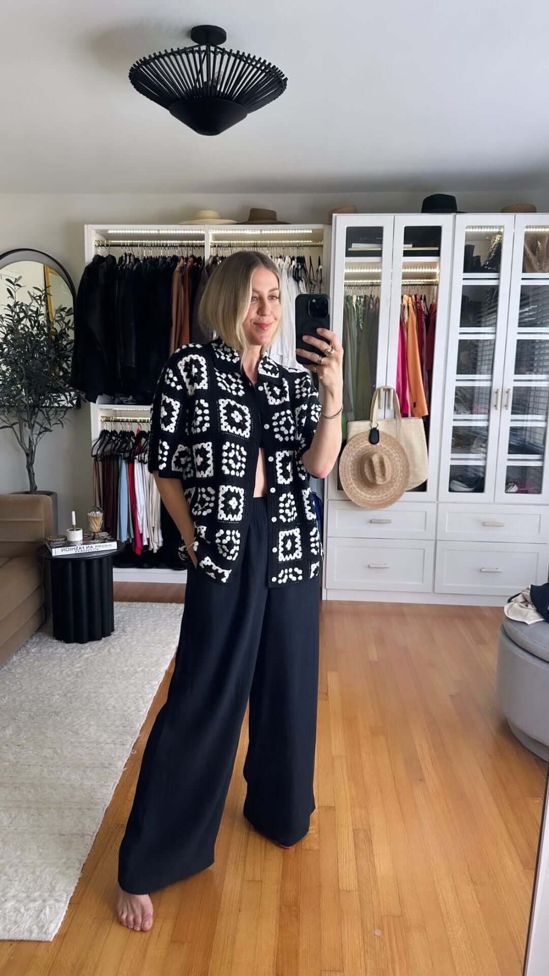 Woman in patterned black and white oversized shirt and wide leg pants, standing in a stylish room with clothing rack.