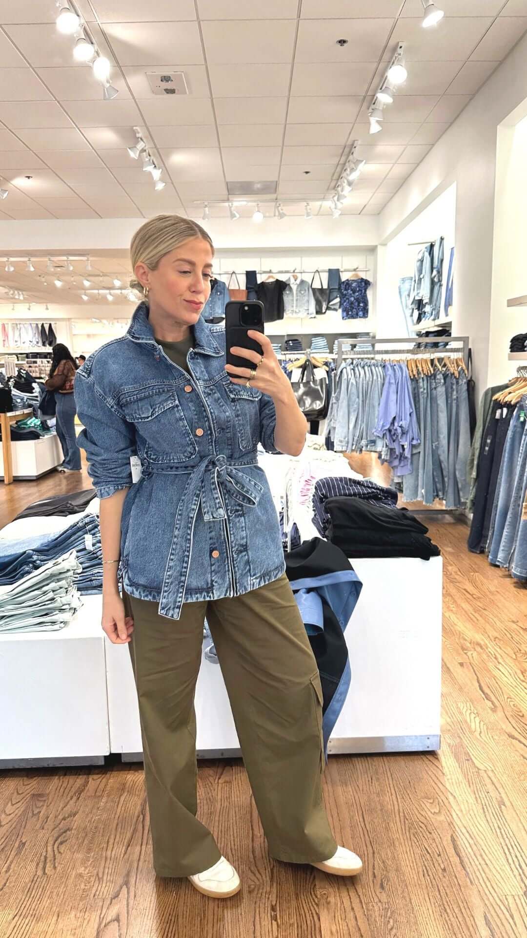 Woman in a denim jacket and green pants poses in a clothing store, showcasing casual fashion style.
