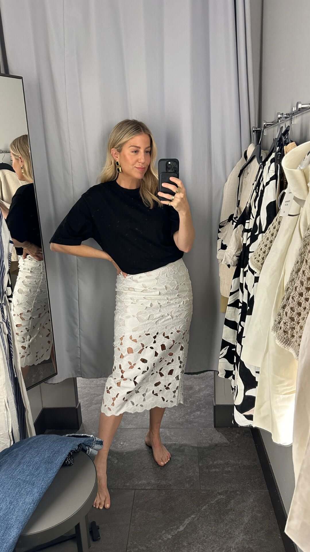 A woman posing in front of a mirror, wearing a black top and a white skirt with cut-out details, in a stylish fitting room.