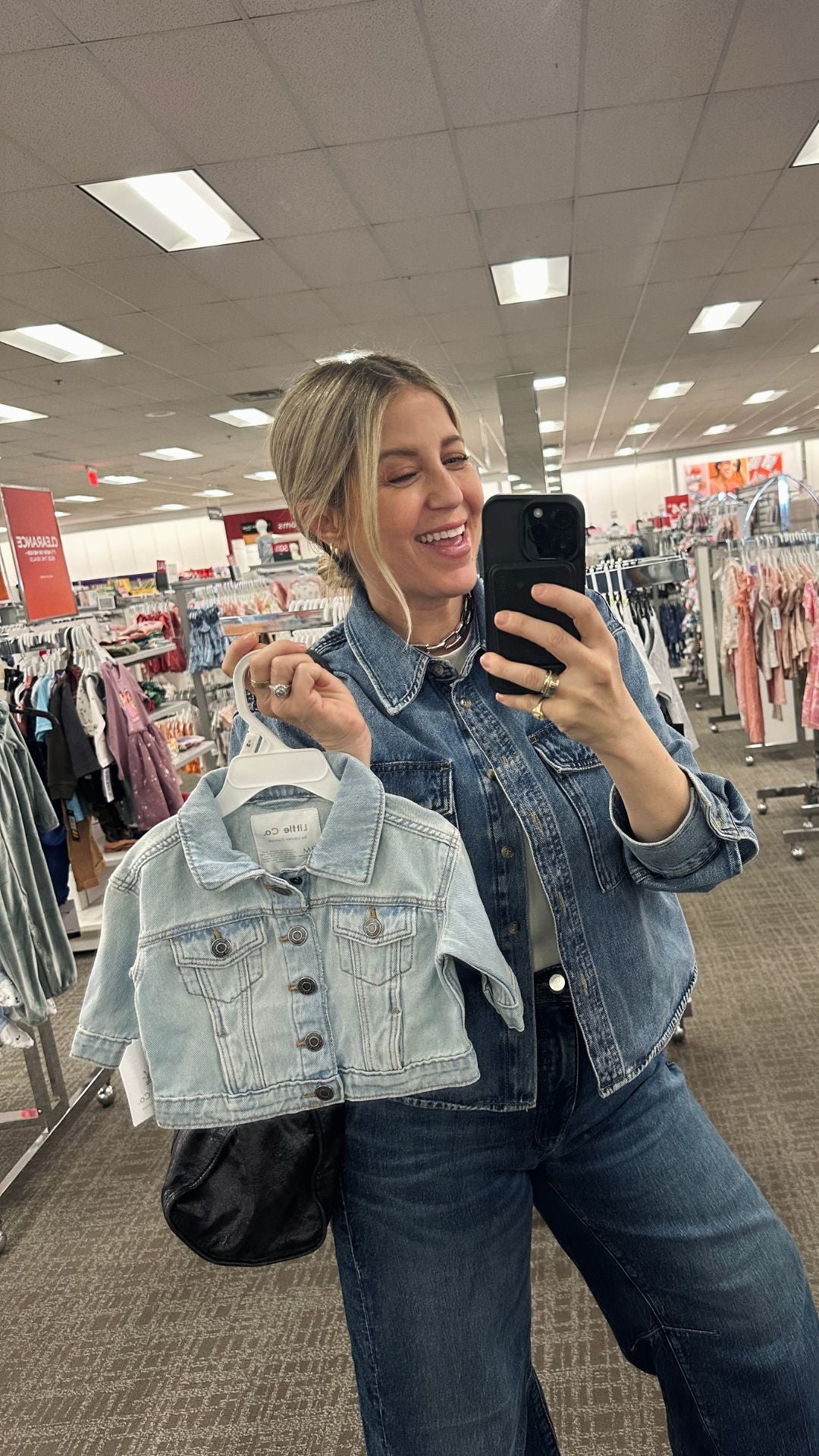 Woman smiling in a denim jacket, holding a small denim jacket in a clothing store, showcasing trendy outfits.