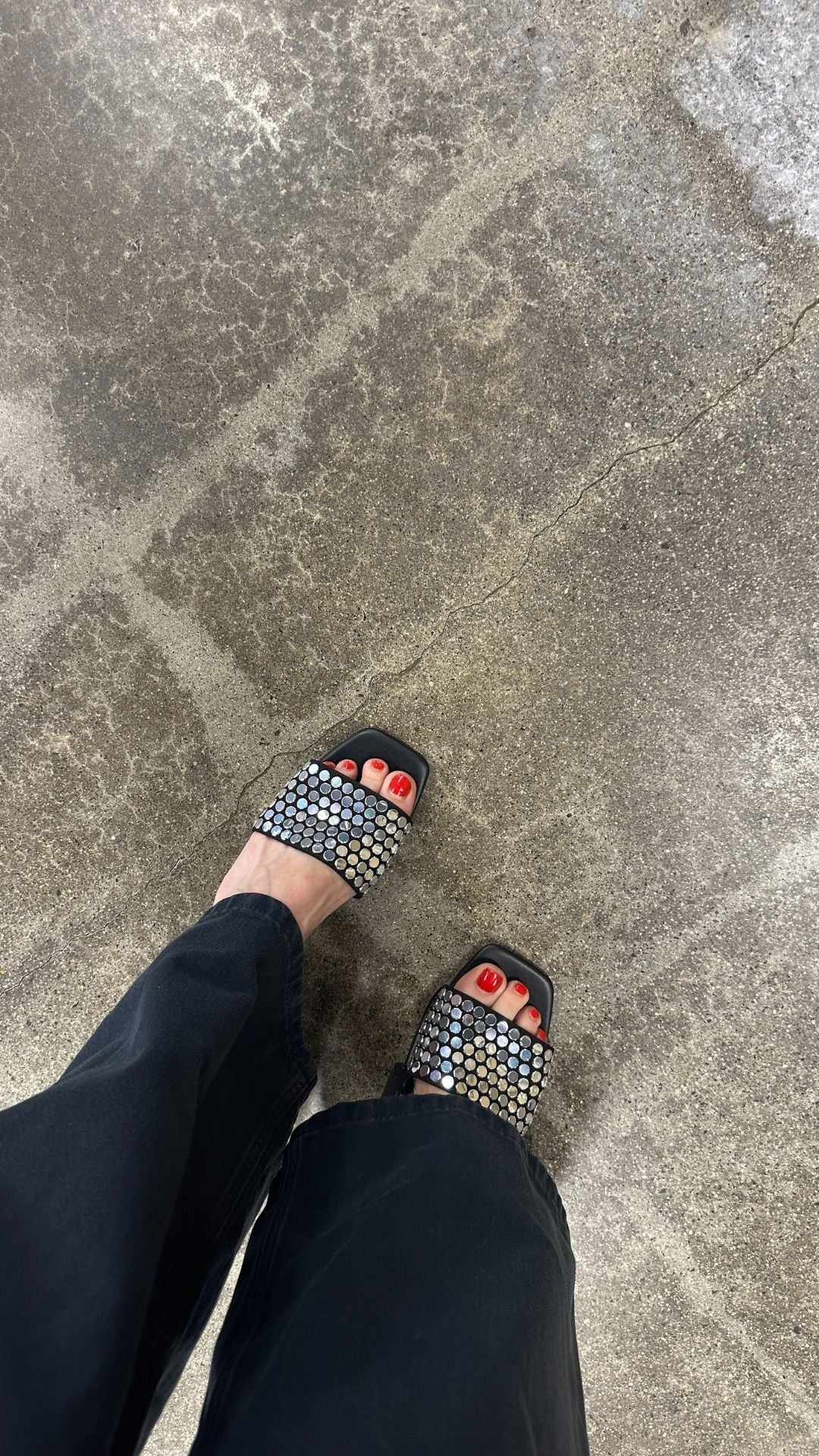 Stylish black sandals with rhinestones and red nail polish on toes, standing on a concrete floor.