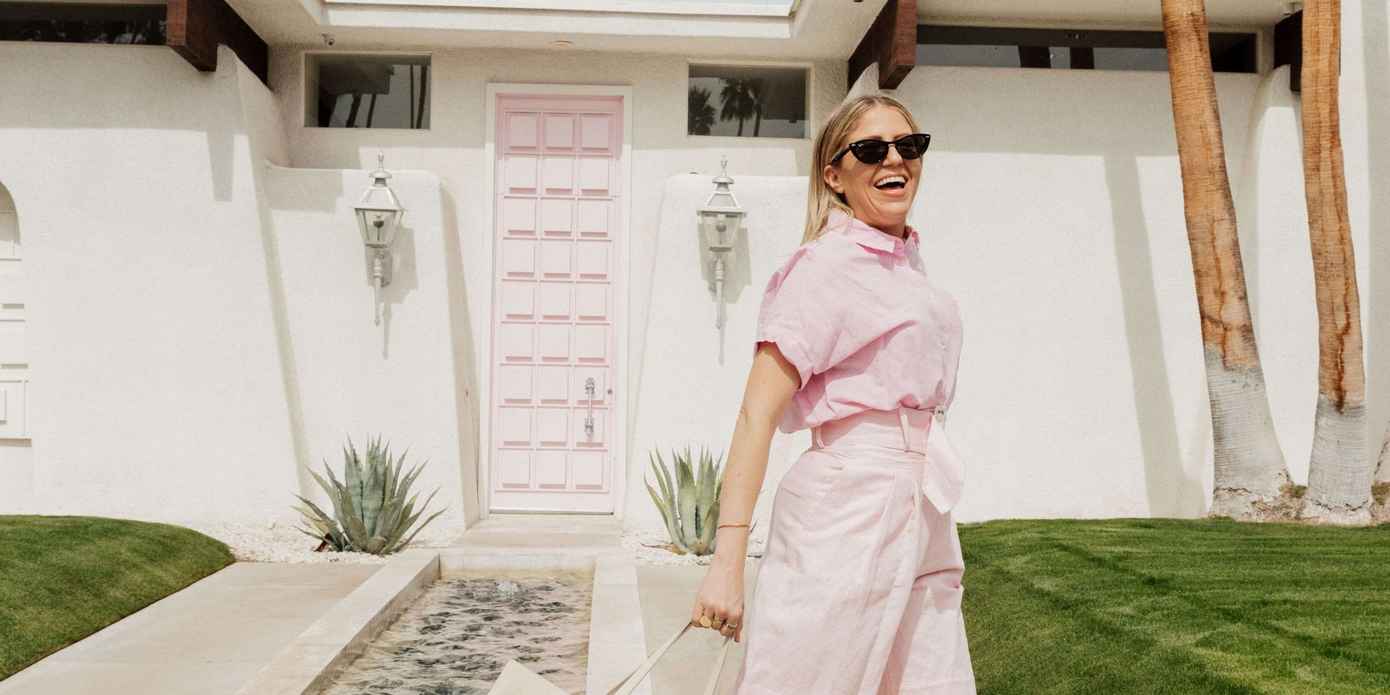 Woman in sunglasses wearing a pink outfit, smiling outside a white house with a pink door.