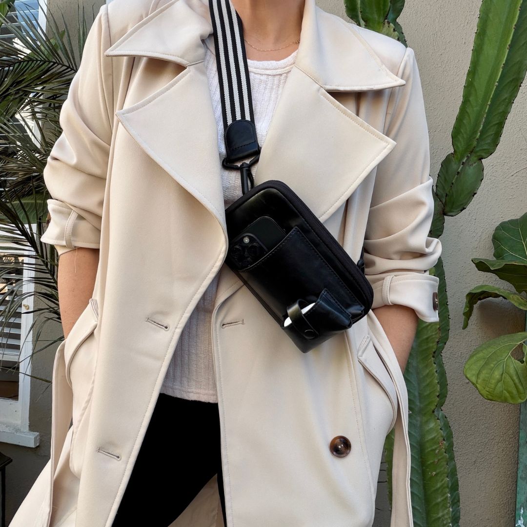Person wearing a beige trench coat with a black crossbody bag, standing outdoors with plants in the background.
