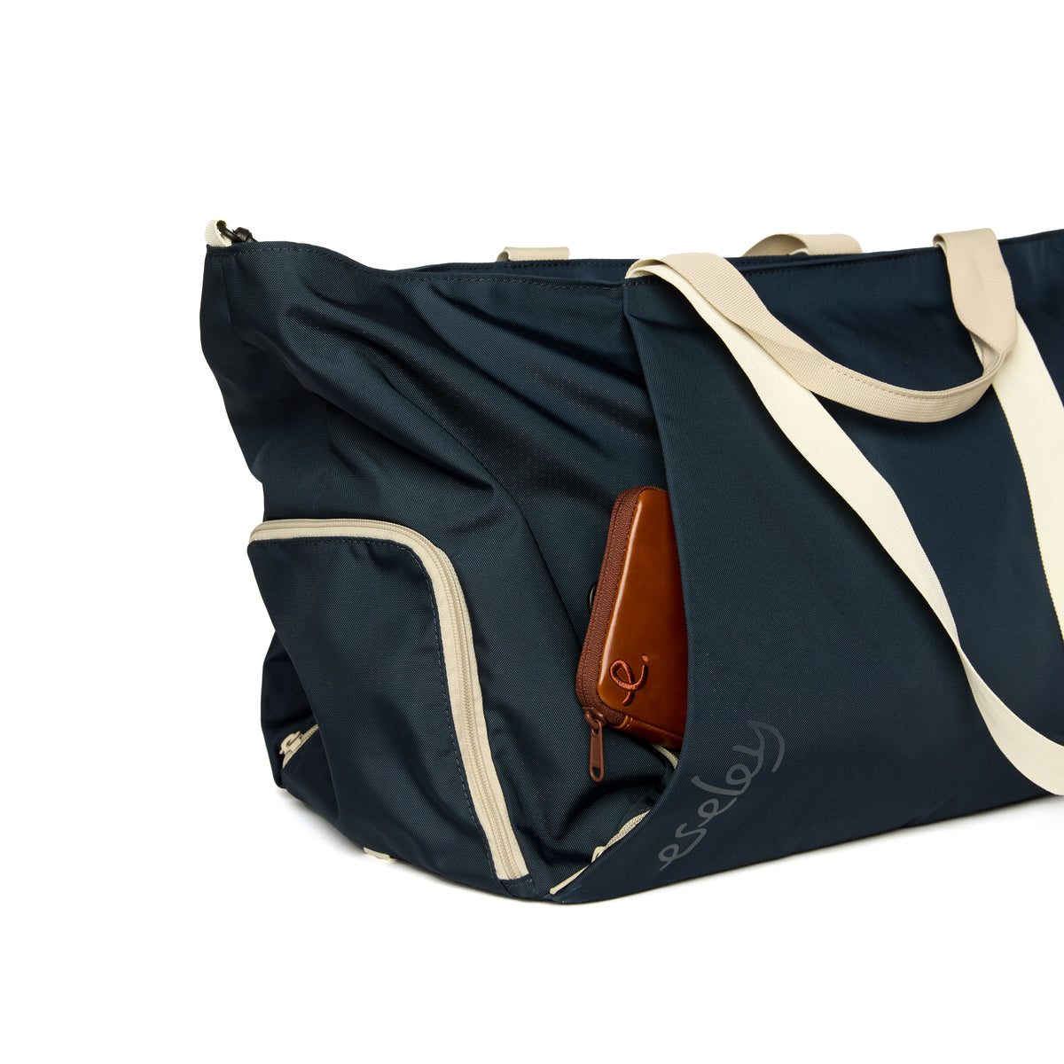 Navy duffel bag with beige straps and a brown leather wallet on a white background