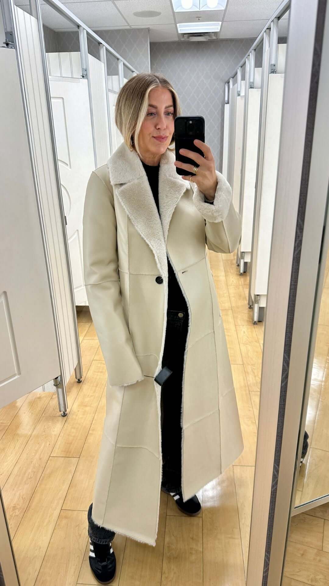Woman in a stylish cream coat taking a mirror selfie in a dressing room.