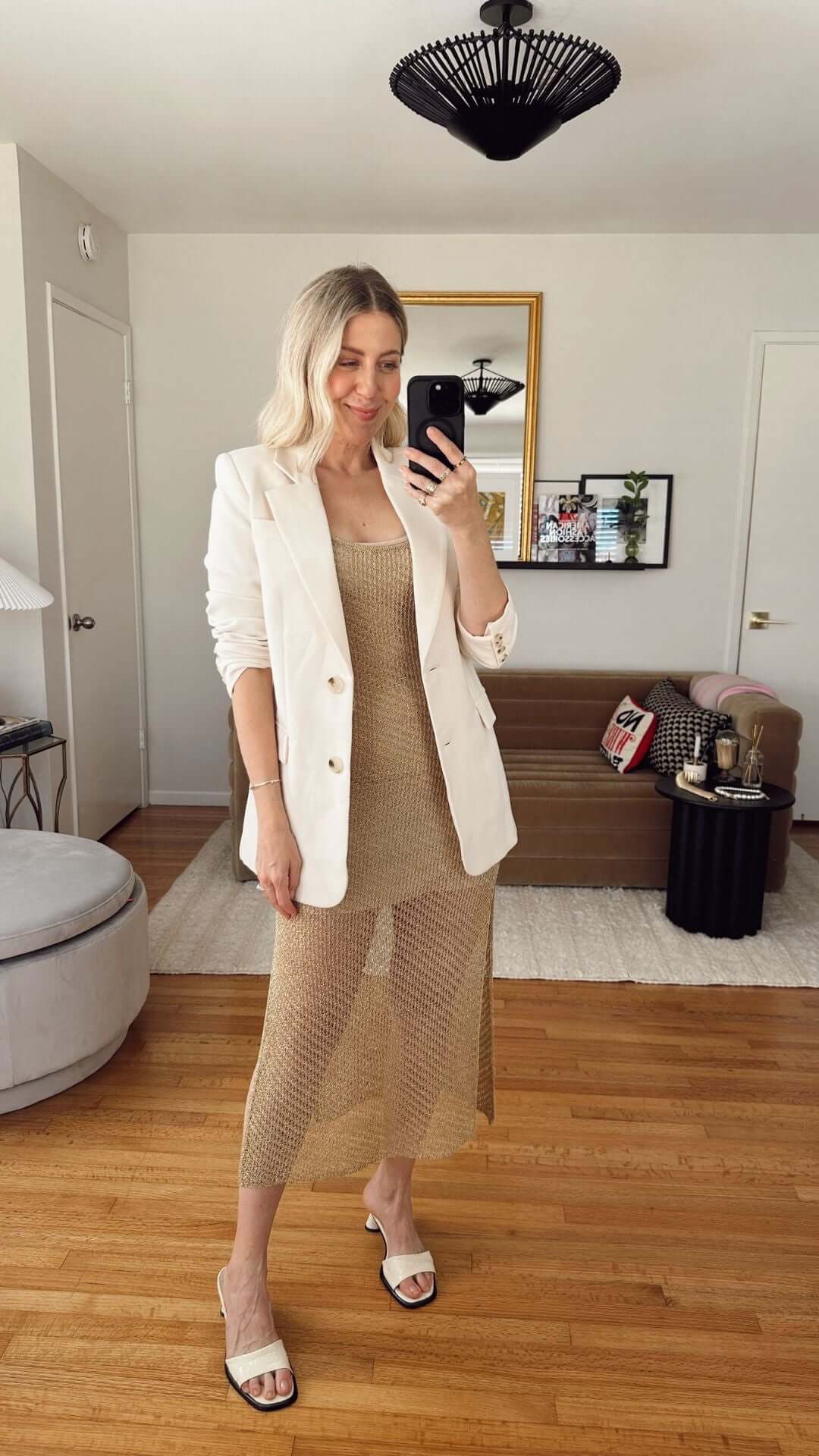 Woman in a chic white blazer and gold dress posing in a stylish living room with modern decor.
