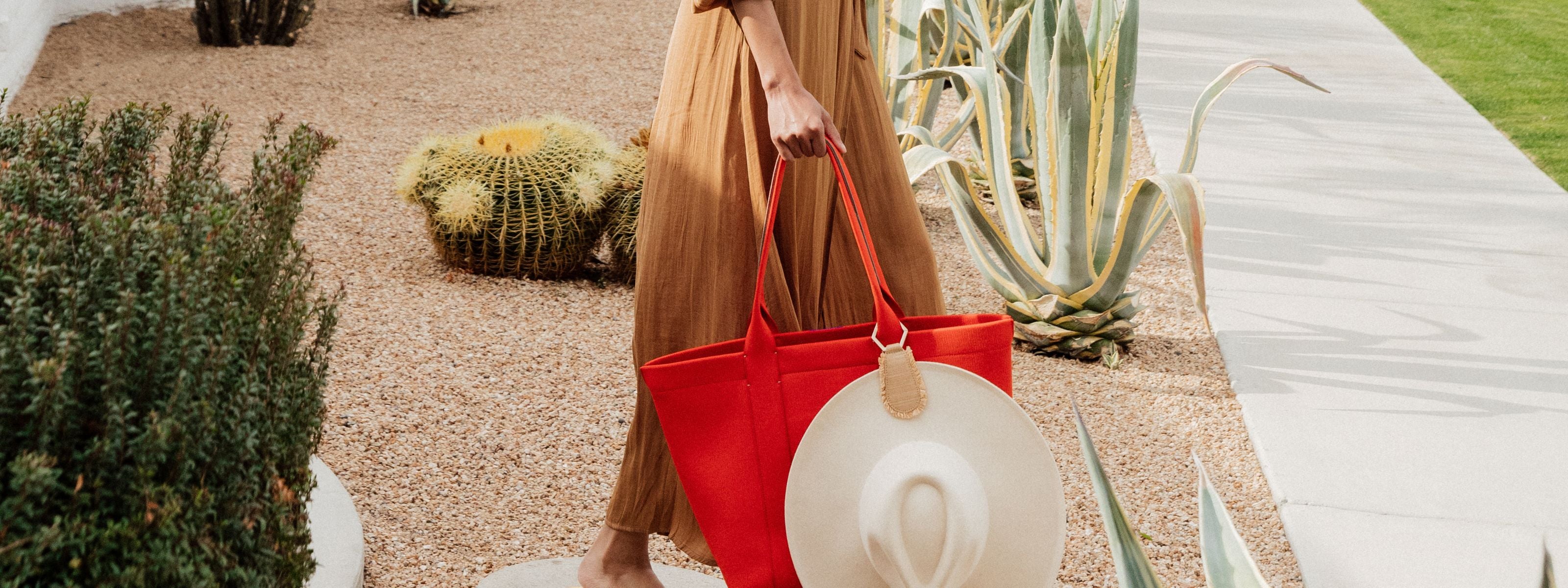Woman in a brown dress carrying a bright red tote bag and a large sun hat in a desert garden setting.