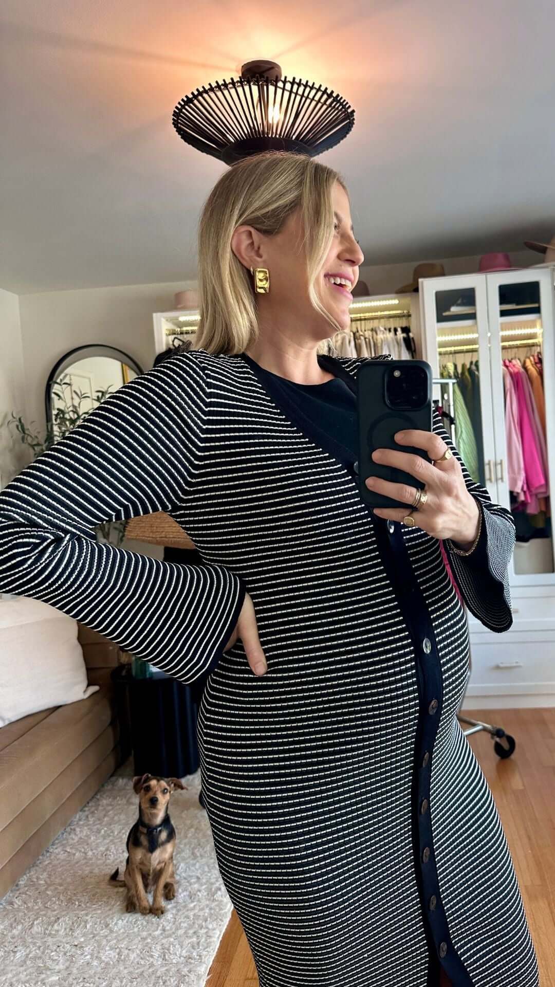 A smiling woman in a striped cardigan poses with a playful hairstyle; a dog is seated nearby.