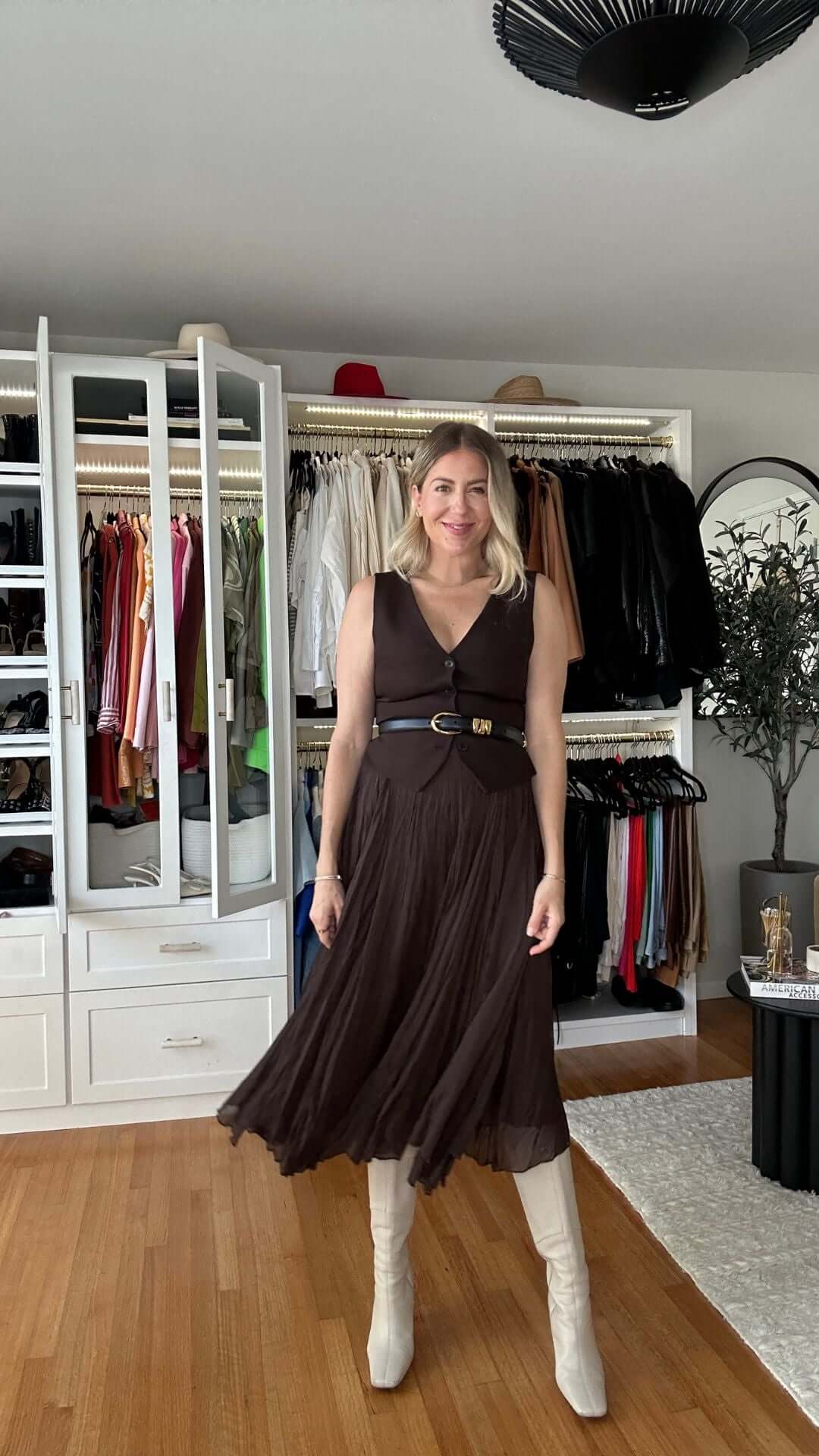 Woman in a brown dress with a belt and white boots standing in a stylish wardrobe room, showcasing fashion and style.