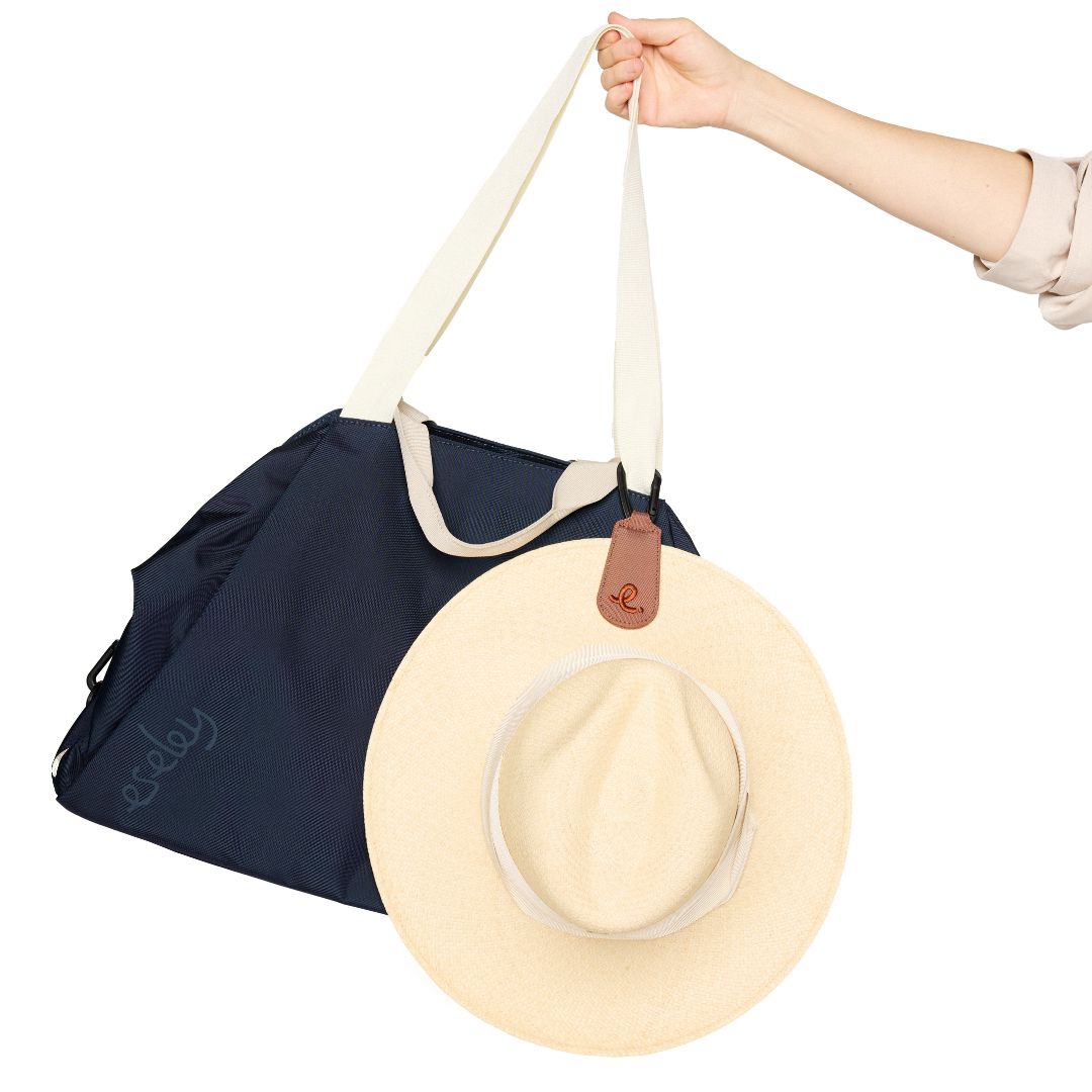 Navy round trip bag with a beige hat hanging on a leather brown hat clip on a white background