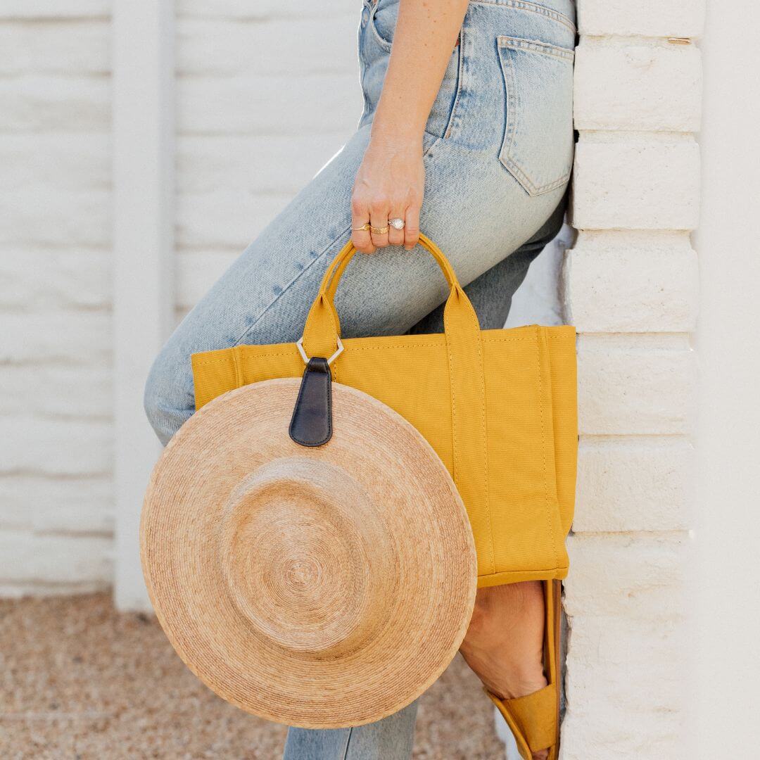 woman in jeans leaning on a wall holding a yellow tote bag with he faux leather drop TOPTOTE hat clip attached with a tan straw hat
