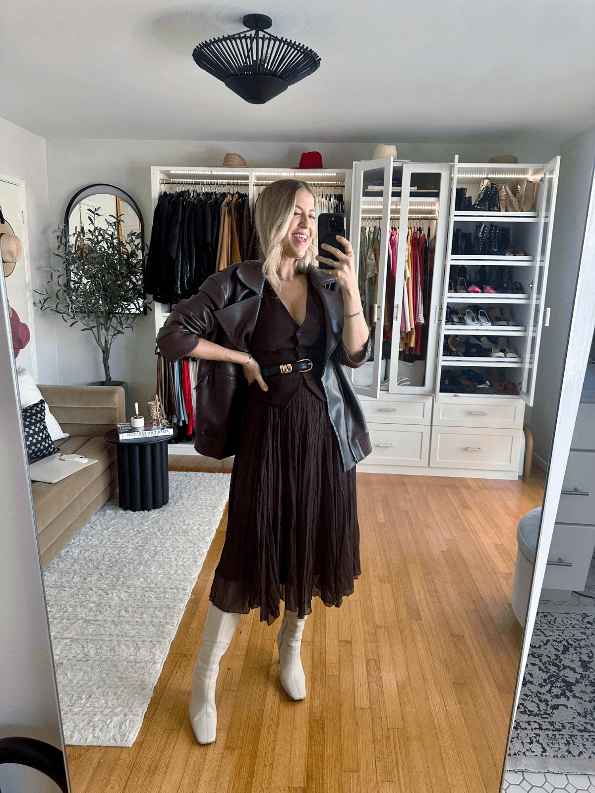 Woman posing in stylish brown dress with a black belt and leather jacket in a chic wardrobe space.