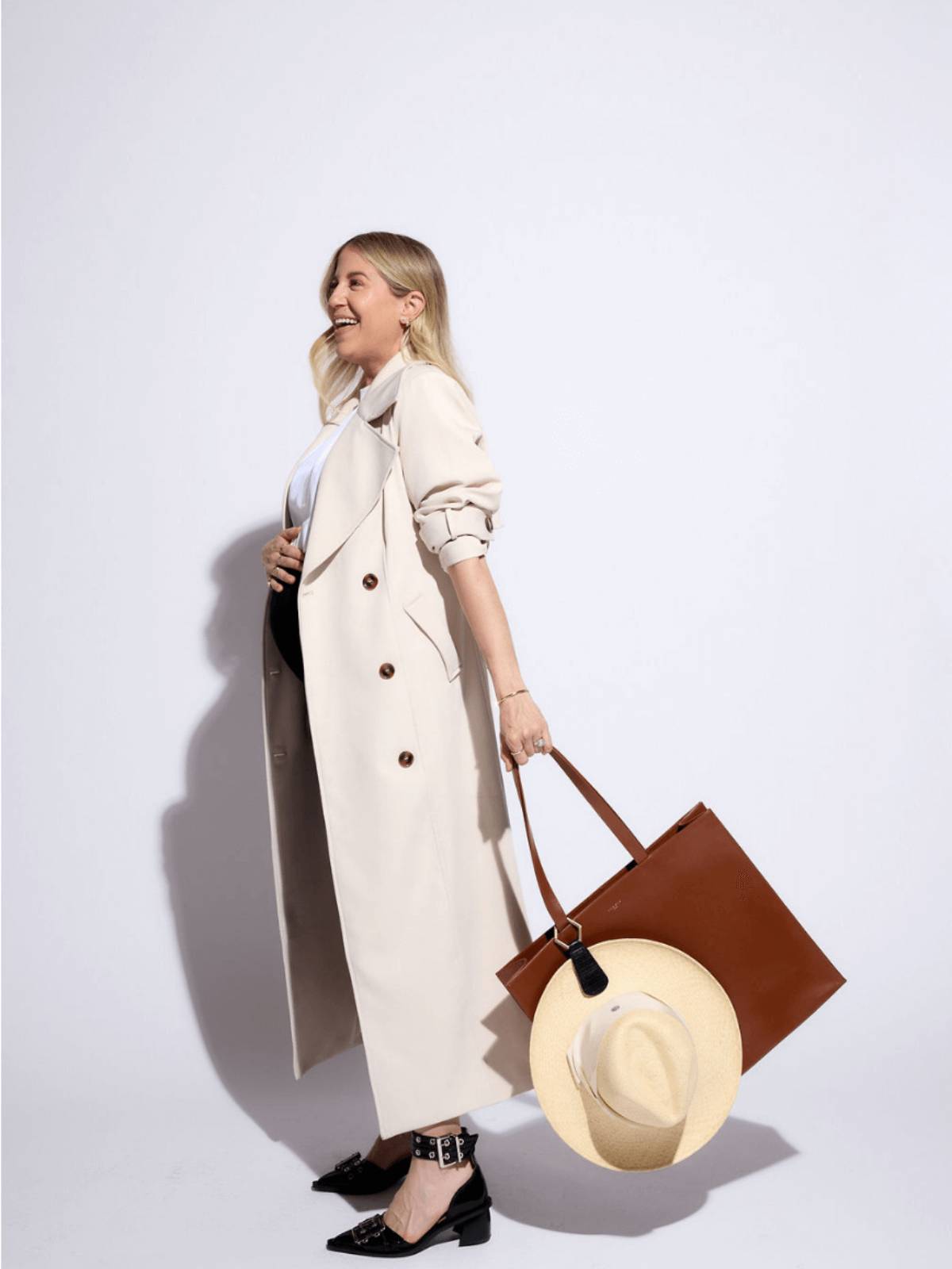 Stylish woman wearing a beige trench coat, holding a brown tote bag and a straw hat, exuding summer elegance.