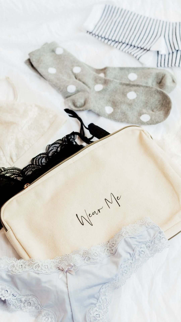 Flat lay of lingerie, socks, and a 'Wear Me' cosmetic bag on a soft background.