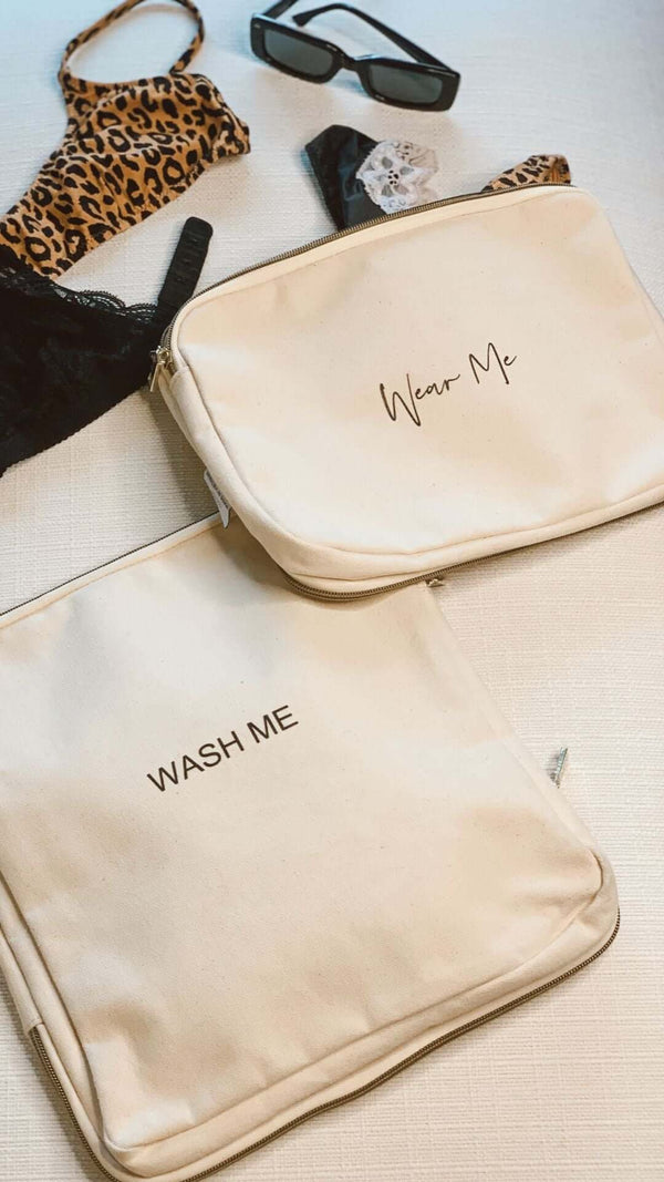 Two travel bags labeled 'Wear Me' and 'WASH ME' next to swimwear and sunglasses on a light surface.