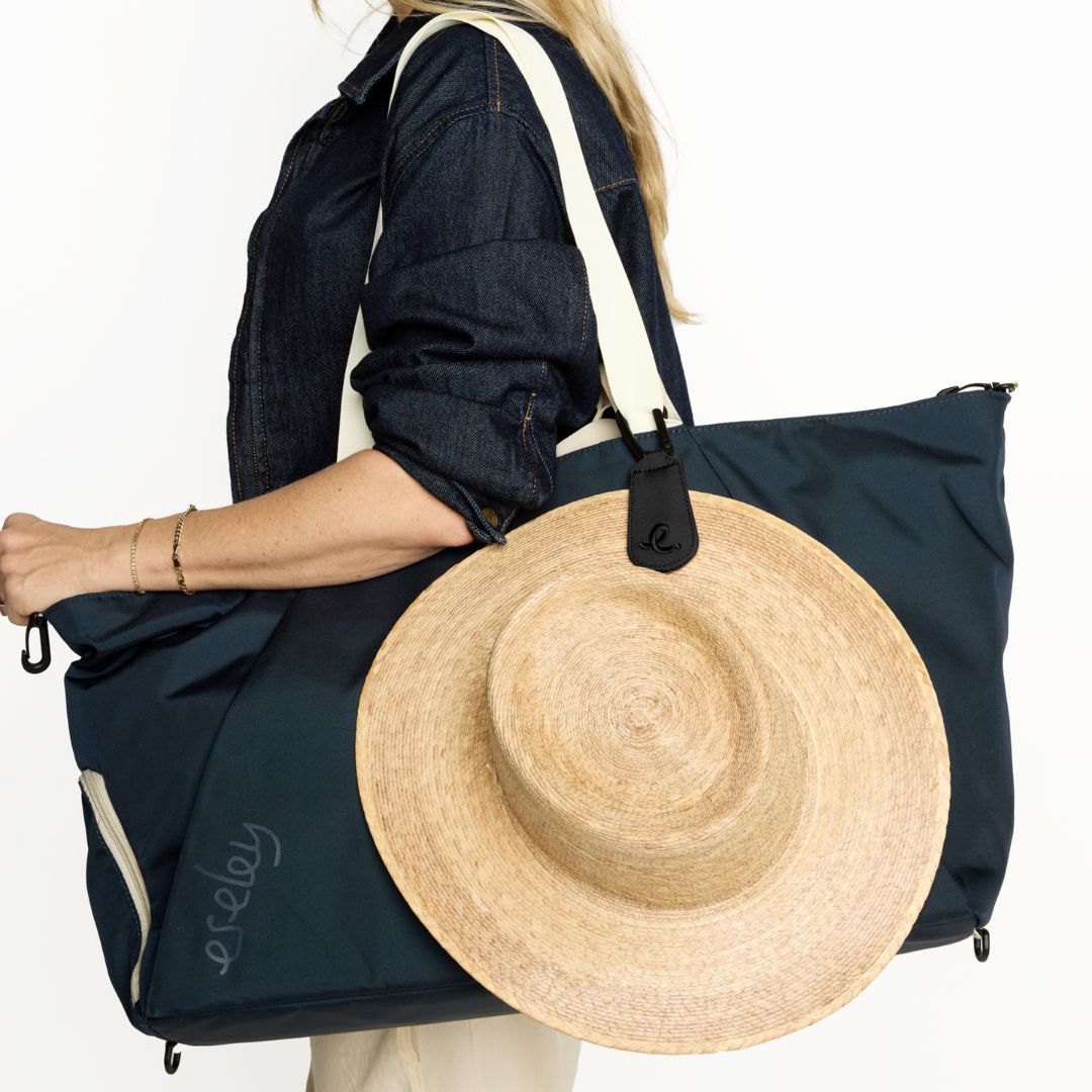 Person holding a navy round trip bag with a black hat clip holding a straw hat on a white background