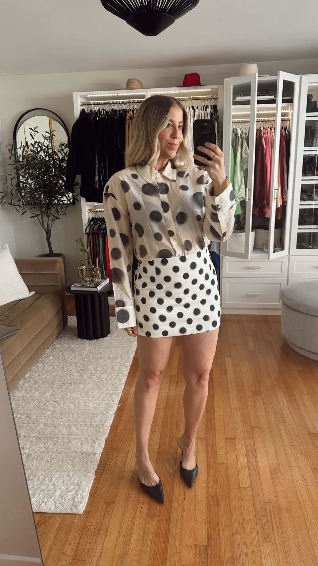 Woman taking a selfie in a polka dot blouse and skirt, standing in a stylish interior with a mirror.