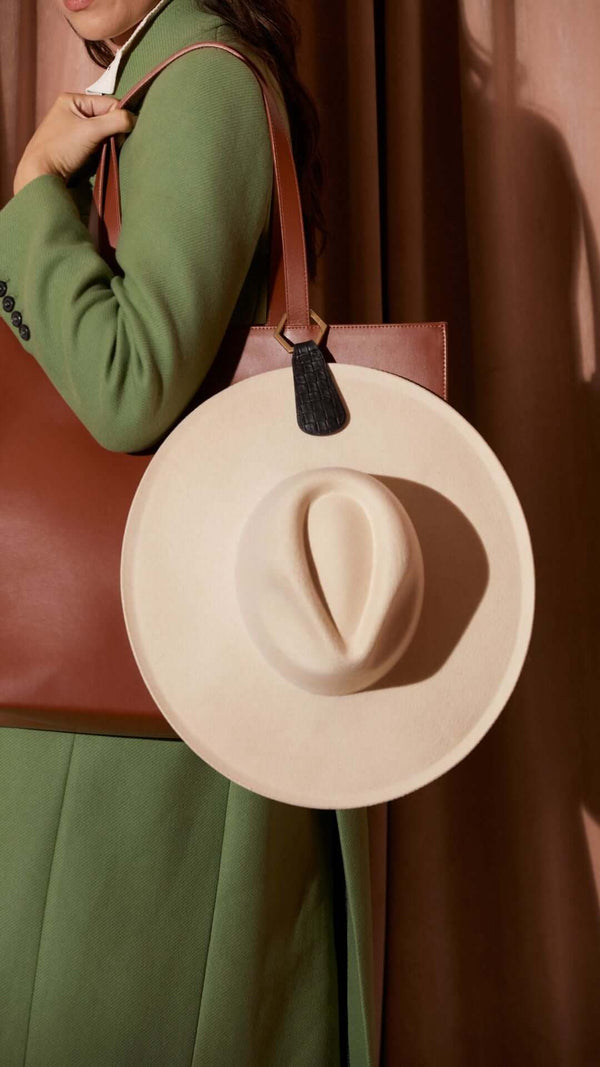 Woman in a green coat holding a brown tote bag with a wide-brimmed cream hat on the side.