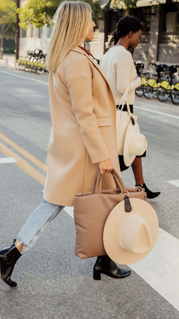 Two women walking on a city street; one in a stylish coat carrying a bag and hat, the other in a cozy sweater.