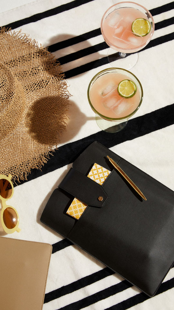 Stylish beach setup with drinks, a straw hat, sunglasses, and a chic black clutch on a striped towel.