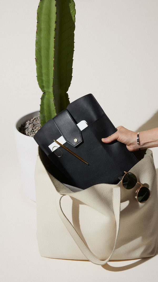 Stylish black folder being taken from a cream tote bag beside a tall cactus and sunglasses.
