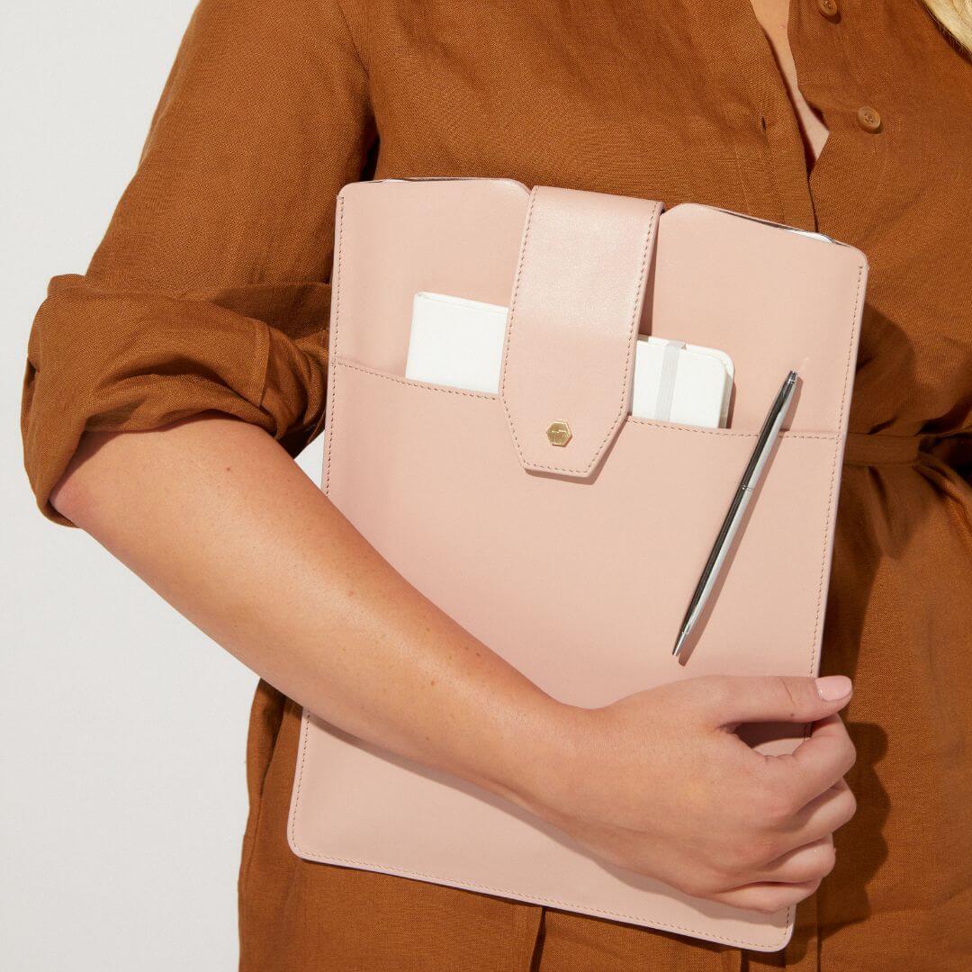 woman holding the Lindsay Albanese blush laptop sleeve with a notebook stuffed in the front pocket with a pen.
