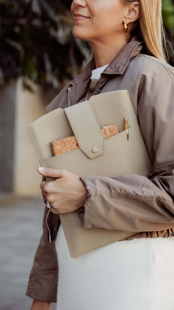 Woman holding a stylish beige folder with a cork notepad and pen, dressed in a chic outfit outdoors.
