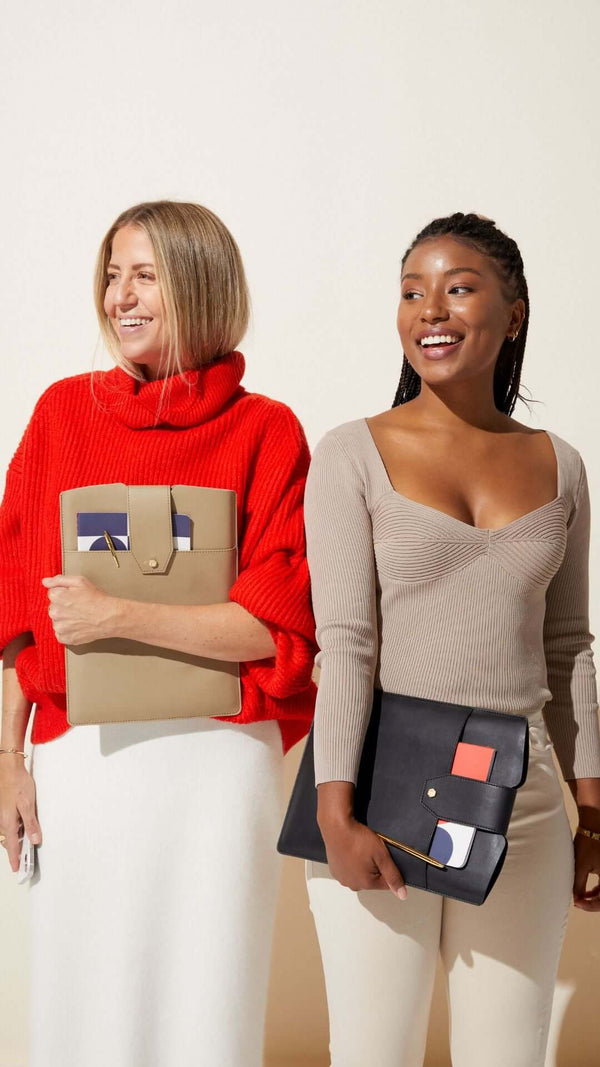 Two women smiling together, one in a red sweater and the other in a tan top, holding stylish bags.