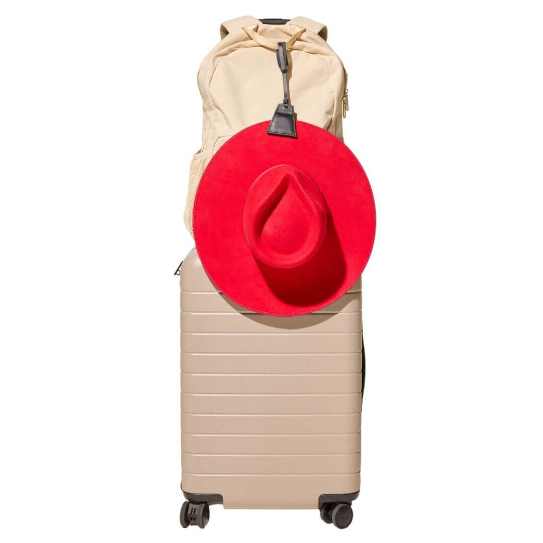 Lasso TOPTOTE hat clip in black holding a red hat and attached to a cream backpack that is resting on a beige carry-on suitcase.
