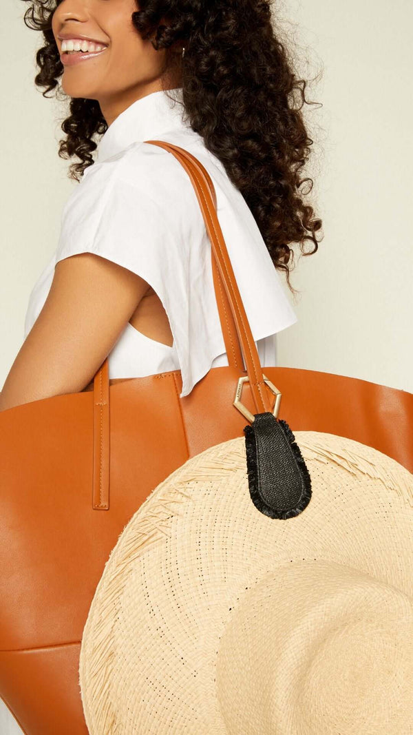 Smiling woman with curly hair carrying a stylish brown tote bag and a wide-brimmed straw hat.