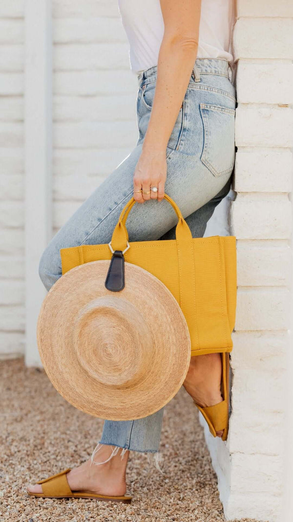 Woman in casual style holding a yellow tote bag and straw hat, standing against a white wall.