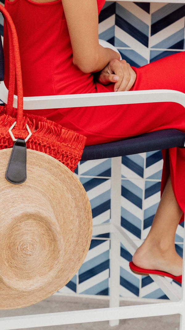 A woman in a red dress sitting on a chair, showcasing a straw hat and a vibrant red bag against a patterned backdrop.
