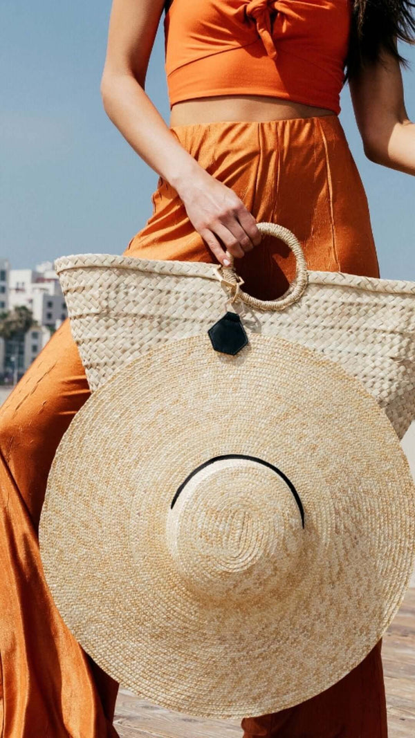 Woman in orange outfit holding a wide-brimmed straw hat and a stylish tote bag by the beach.