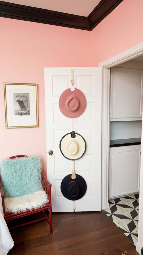 Stylish hats hanging on a door in a pastel pink room, featuring a cozy chair and modern flooring.